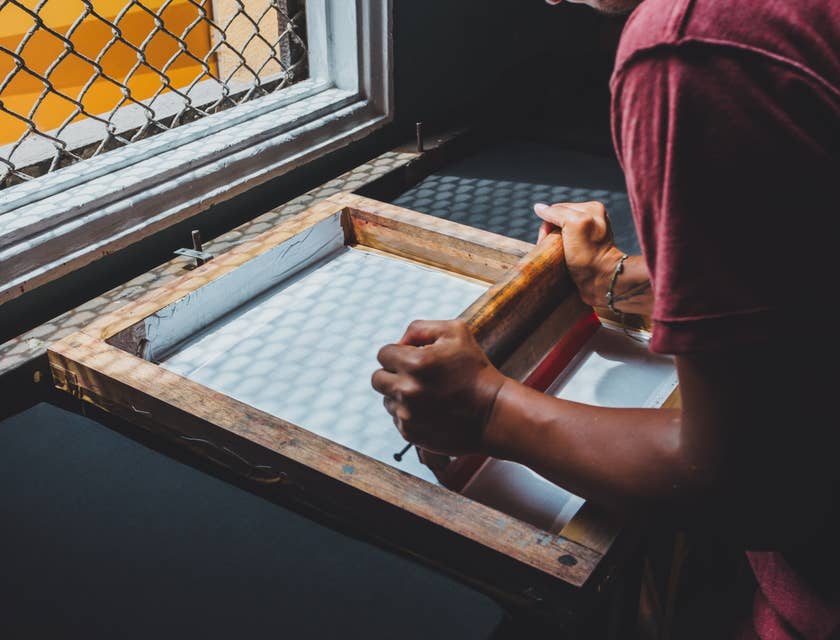 Ein Mann setzt auf einer Werkbank vor einem Fenster ein traditionelles Siebdruckverfahren in einer Druckerei ein.